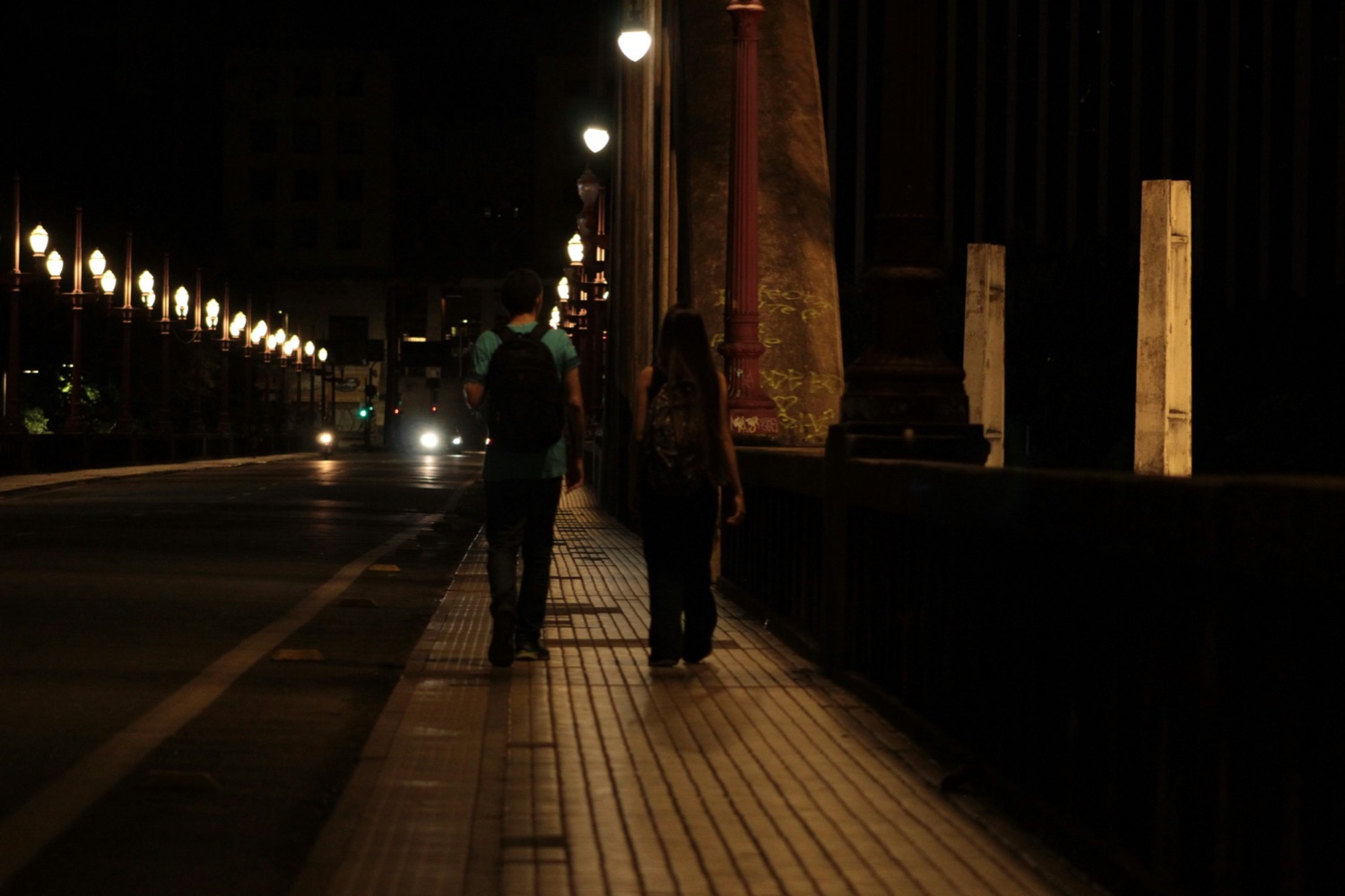 Two people walking along a lamp-lit bridge at night